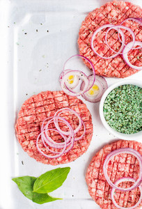 Three raw ground beef meat cutlets for making burgers with onion rings and spices on white wooden background top view