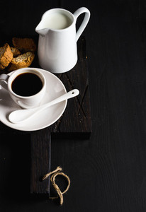 Cup of hot espresso creamer with milk and cookies on dark rustic wooden board over black background copy space