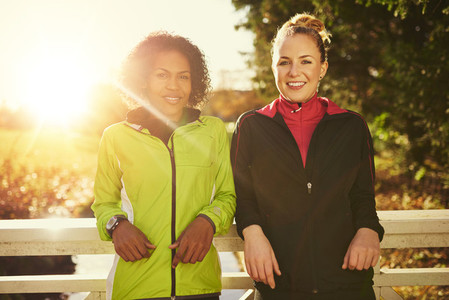 Two smiling sportswomen leaning on bridge and looking at camera