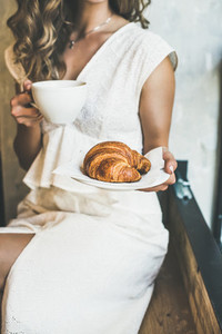 Blond woman holding fresh croissant and cup of cappuccino