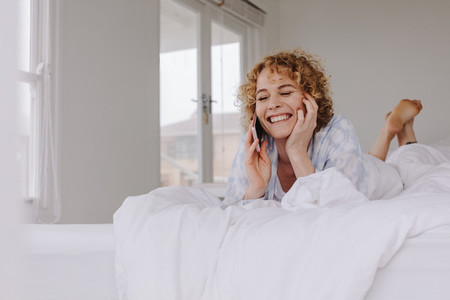 Happy woman lying on bed talking over cell phone