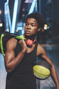 Portrait of young afro man with a basket ball wearing red headphones and a bag waiting friends in urban scenery at dusk