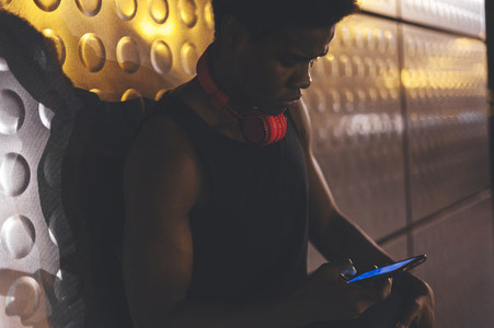 Portrait of young afro man chatting by phone leaning in a wall  wearing sportswear and red headphones in urban scenery at night with city lights
