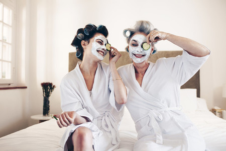 Happy women sitting on bed in bathrobes putting on beauty face m