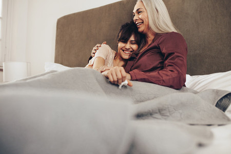 Smiling mother and daughter sitting on bed together covered in b