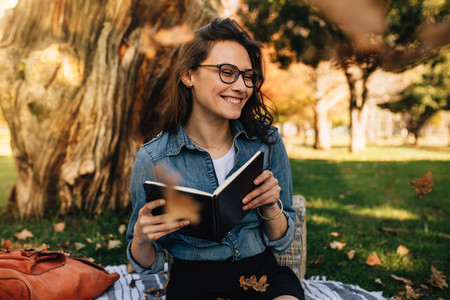 Smiling woman sitting at park with book and leaves falling aroun