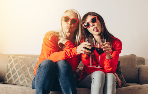 Two women sitting on couch with glass of wine making faces