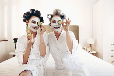 Two women in bathrobes with beauty face pack sitting on bed