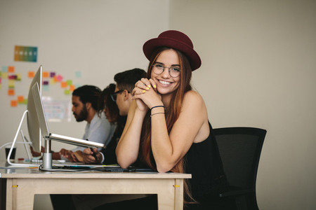 Smiling businesswoman in hat and eyeglasses sitting at her desk