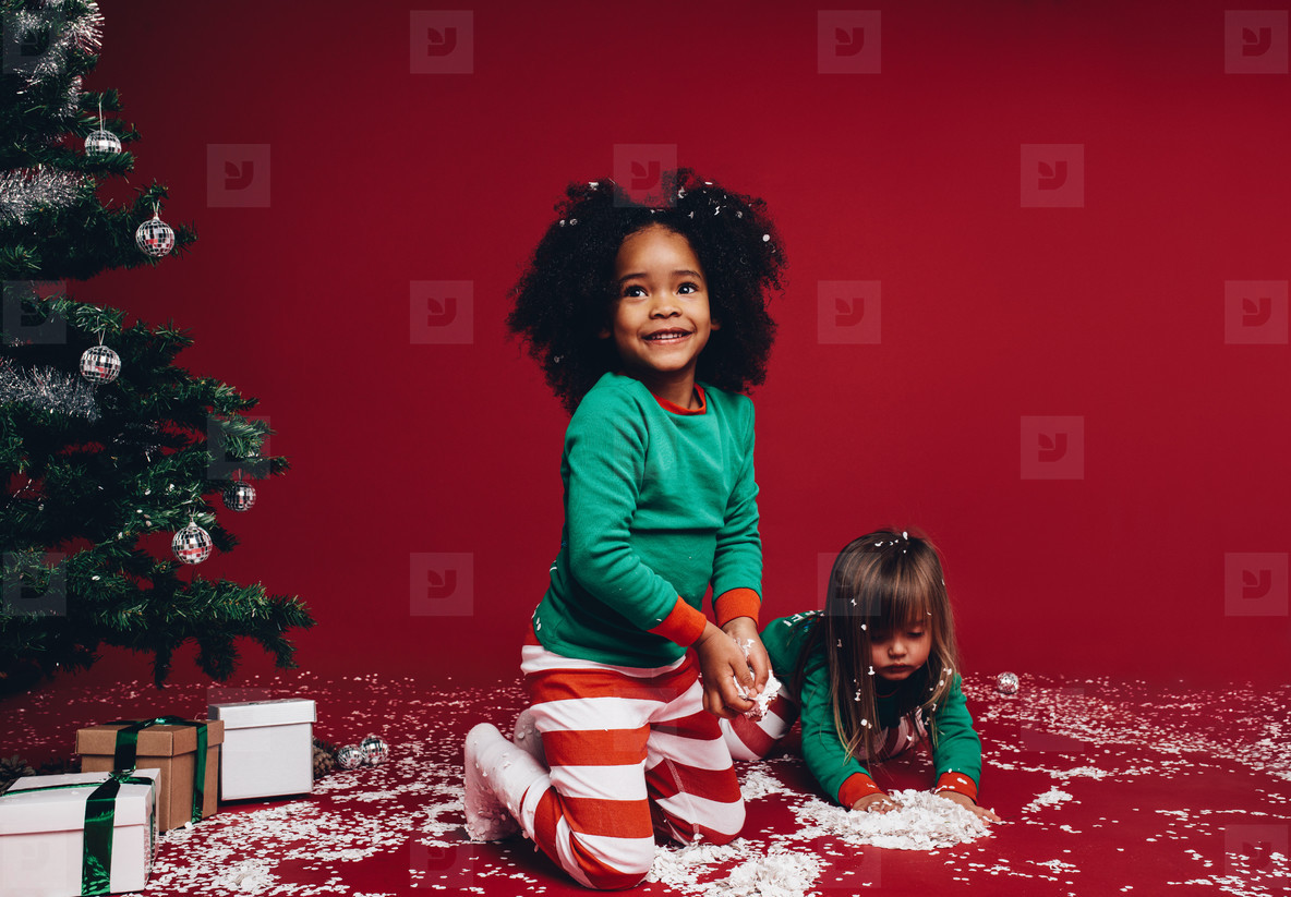 Two kids playing with artificial snow beside a christmas tree