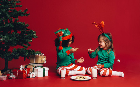 Kids eating cookies sitting beside a christmas tree