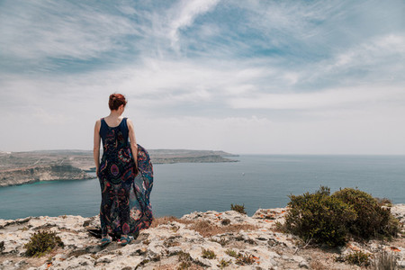 Young redhead woman from the edge of cliff wearing a long dress moving with the wind