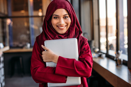 Cheerful islamic woman in hijab with laptop at cafe
