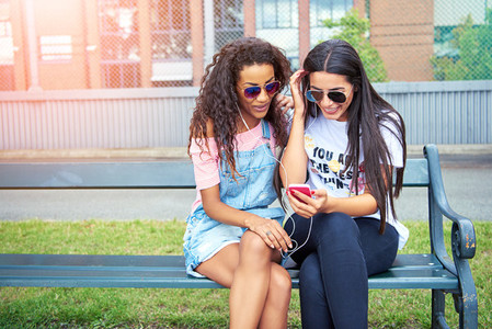 Two smiling girlfriends sitting outside listening to music on earphones