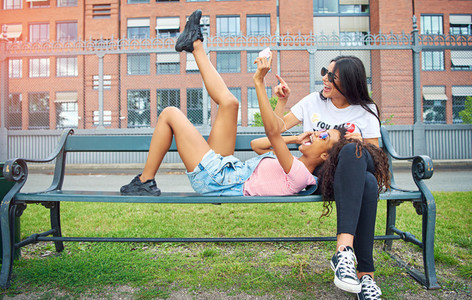 Carefree young girlfriends sitting on a bench outside taking selfies