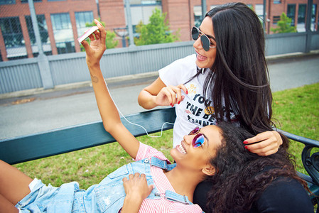Laughing young girlfriends taking selfies on bench outside