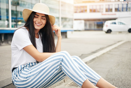 Woman in straw hat and striped pants sits on curb