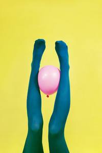 Green legs of a woman holding a pink balloon against a yellow wa