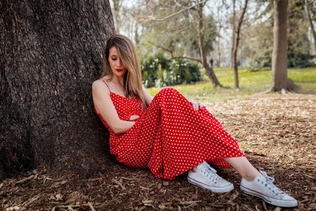 Young serious blond woman sitting near tree with red long dress