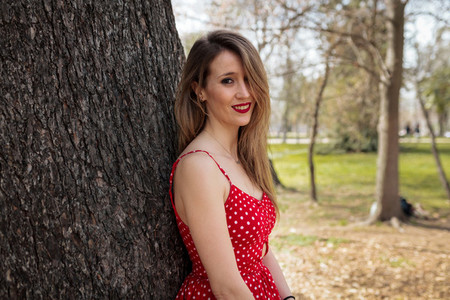 Young blond woman with red dress leaning against a tree