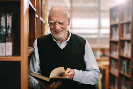 Smiling senior man looking at a book standing in a library