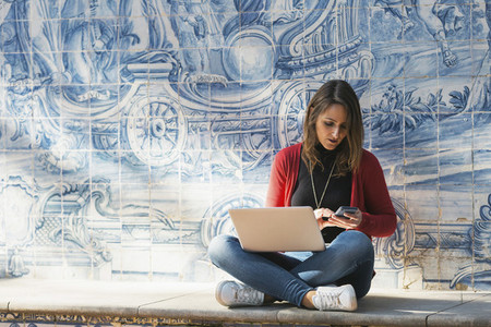 Woman using laptop and smart phone against mosaic wall 01