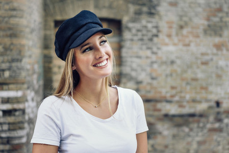 Young blonde woman wearing cap smiling near a brick wall