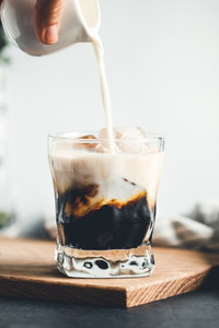 Pouring milk in a glass with espresso and iced cubes in a modern kitchen in the morning