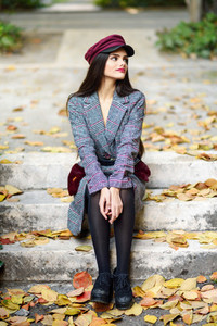 Young beautiful girl wearing winter coat and cap sitting on steps full of autumn leaves