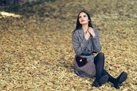 Beautiful girl wearing winter coat sitting on the floor of an urban park full of autumn leaves