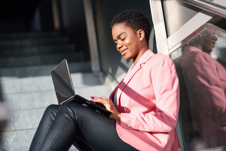 Smiling black businesswoman sitting on urban steps working with a laptop computer