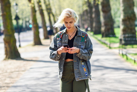 Young urban woman with modern hairstyle using smartphone walking in street in an urban park