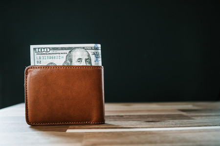 One hundred dollar bill in a brown leather wallet on a rustic table with a black background  Copy space