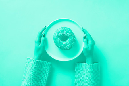 Female hands holding donut on plate over mint color background  Trendy green and turquoise color  Top view  flat lay  Sweet  dessert  diet concept  Weight lost after holidays
