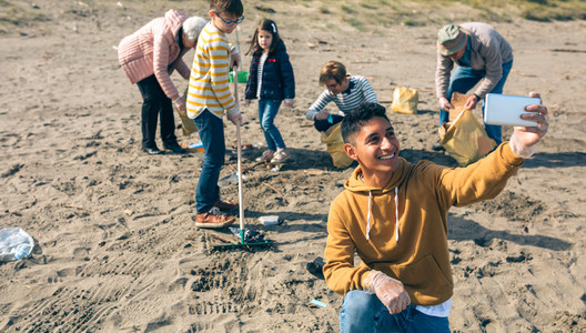 Young man taking selfie to group of volunteers cleaning beach