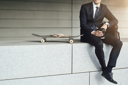 Stylish man wearing suit sitting near skateboard