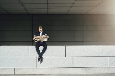 Businessman sitting on brickwall and reading newspaper