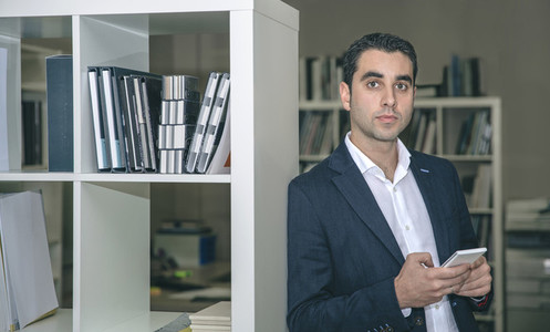 Businessman holding smartphone leaning on a bookcase