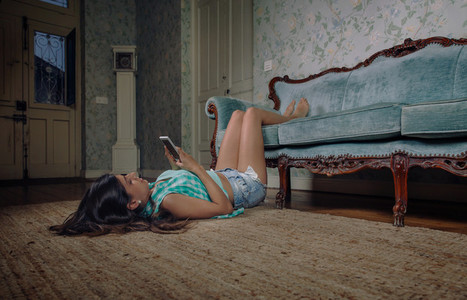 Woman looking smartphone lying on carpet with legs over sofa