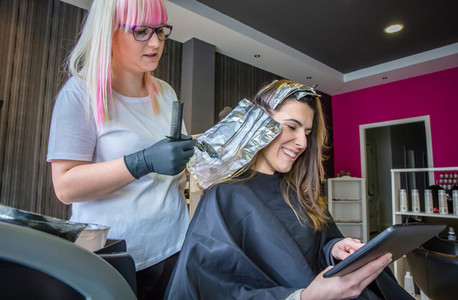 Hairdresser applying hair dye to woman looking digital tablet
