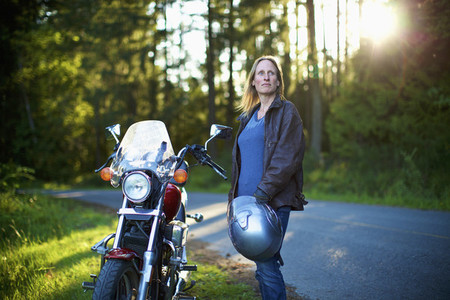 Confident woman standing at motorcycle at roadside in woods