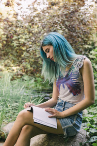 Young woman with blue hair writing in journal in park