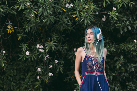 Young woman with blue hair listening to music with headphones in front of flowering tree