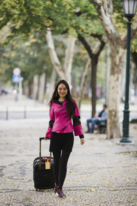 Smiling female tourist pulling suitcase along treelined sidewalk