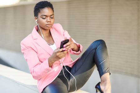 Black businesswoman sitting outdoors using smartphone with earphones