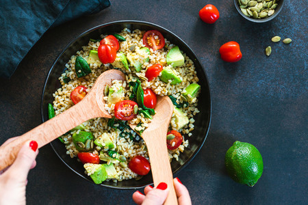 Hands mixing healthy salad with bulgur avocado spinach and cherry tomatoes Top view