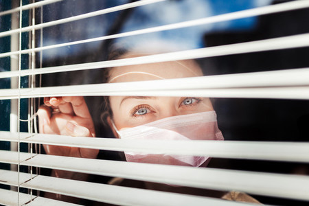 Woman looking out the blind window wearing a surgical mask