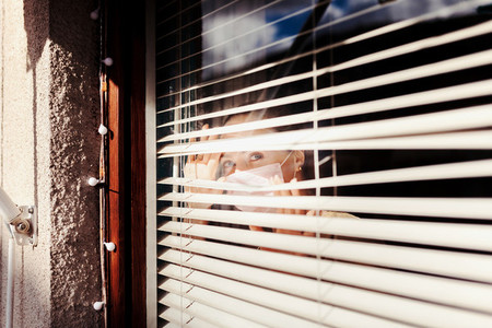 Woman looking out the blind window wearing a surgical mask
