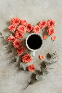 Cup of black coffee surrounded with fresh coral roses and eucalyptus branches on a textured beige background