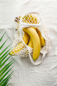 Bananas in a net produce bag on a linen napkin decorated with palm leaf  Eco friendly and summer concept